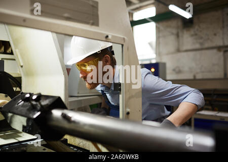 Serious engineer in hardhat and protective glasses watching for the work of machine in the factory Stock Photo