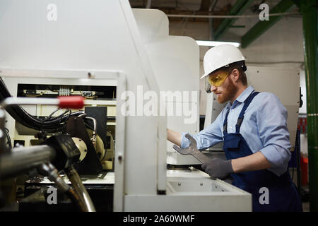 Young bearded repairman in protective workwear using wrench and working on lathe in the plant Stock Photo