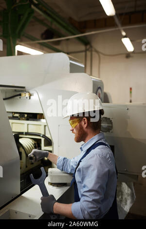 Bearded young man in work wear and protective glasses holding wrench and repairing the lathe Stock Photo