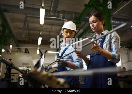Female foreman in work wear using digital tablet and controlling the work while man working with iron pipes in the plant Stock Photo