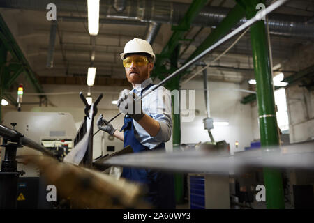 Manual worker in protective workwear and glasses wearing gloves holding iron pipe while working in the factory Stock Photo