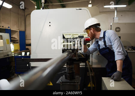 Young operator in protective glasses and workwear standing and working on lathe in the factory Stock Photo