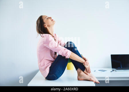 Young woman sitting on desk in office with eyes closed and relaxing Stock Photo
