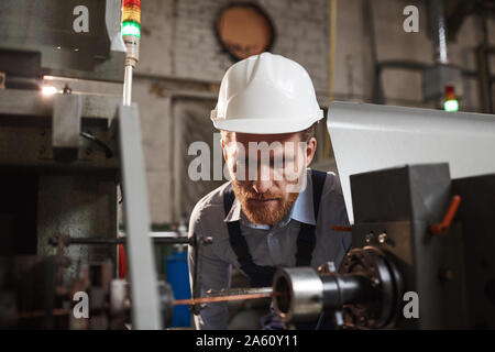 Young mechanic in hardhat looking at the work of machine and controlling the process in the factory Stock Photo