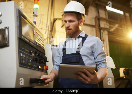 Engineer in protective work wear working with machine and using digital tablet in the plant Stock Photo