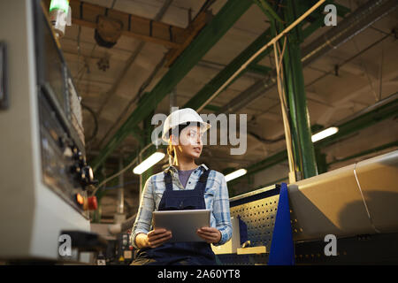 Female technician in work helmet controlling the work of machine with digital tablet in the plant Stock Photo