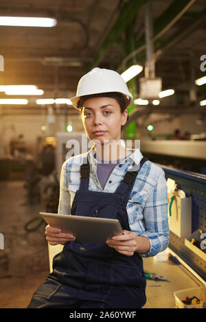 Portrait of female engineer in overalls and in hardhat looking at camera while using digital tablet in the plant Stock Photo