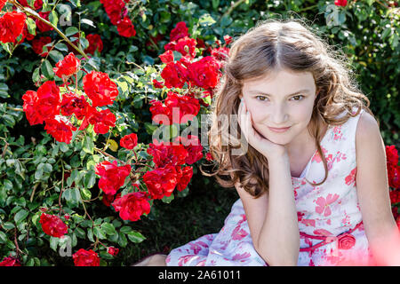 Portrait of girl wearing summerdress with floral design sitting on a meadow beside red rosebush Stock Photo