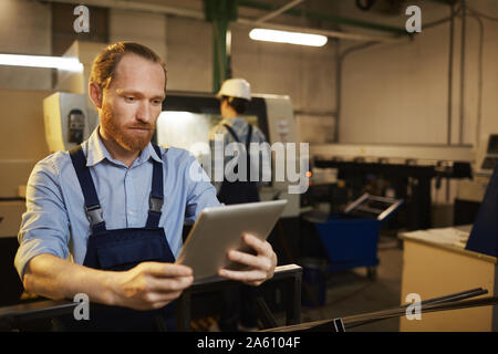 Young bearded engineer in overalls standing and working on digital tablet with woman working in the background in factory Stock Photo