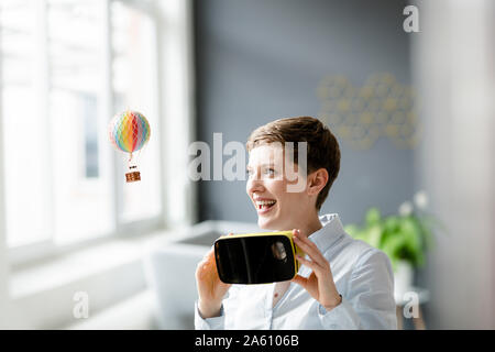Smiling woman with VR glasses and small hot-air balloon in office Stock Photo