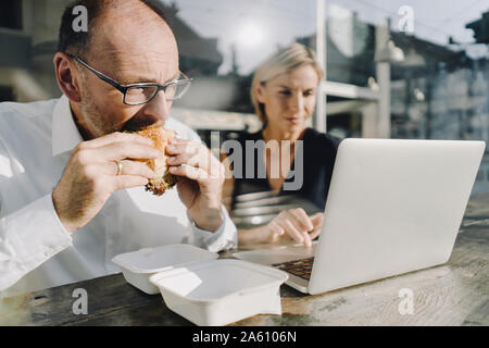Businessman eating hamburger in coffee shop, while colleague is using laptop Stock Photo