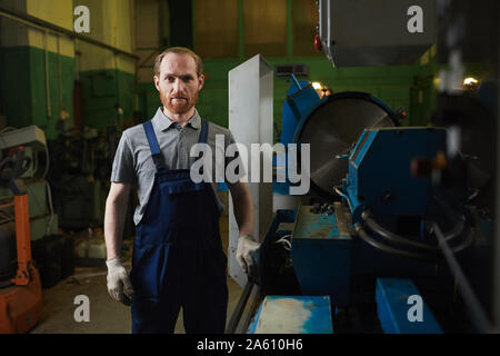 Portrait of young bearded mechanic in overalls looking at camera while standing in the factory Stock Photo