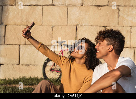 Young couple taking selfies with smartphone, sitting in front of wall Stock Photo