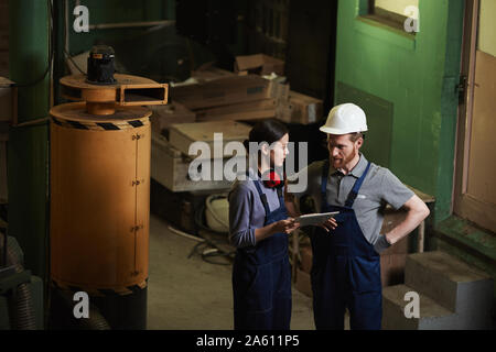 Two manual workers in work wear using digital tablet and talking to each other while standing in the plant Stock Photo