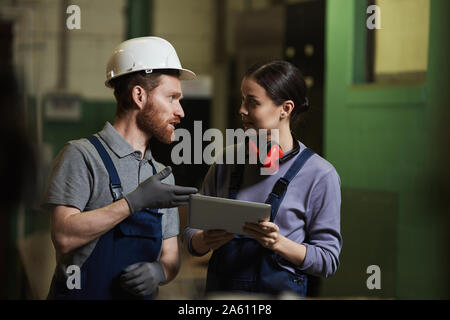 Manual worker in overalls and in work helmet discussing work with female worker and they using digital tablet in the factory Stock Photo