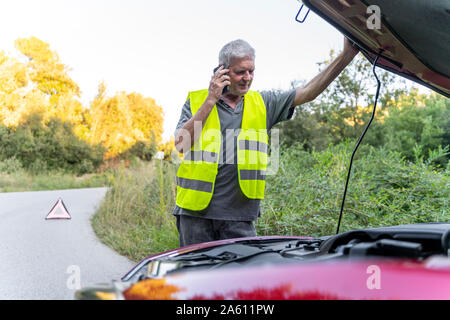 Senior man standing at his broken car wearing a safety vest and using his smartphone Stock Photo
