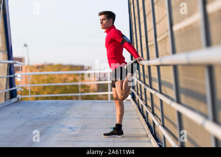 Jogger with smartphone in arm pocket, stretching his leg on a bridge railing Stock Photo