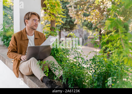 Portrait of pensive man sitting on wall of bower working on laptop Stock Photo