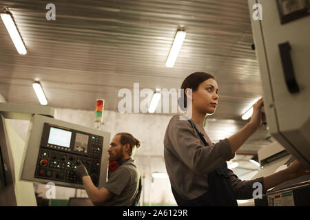 Serious woman in work wear standing and controlling the work of machine with her colleague in the background Stock Photo