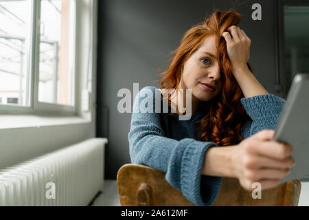 Portrait of redheaded woman with digital tablet sitting in a loft leaning on back rest Stock Photo