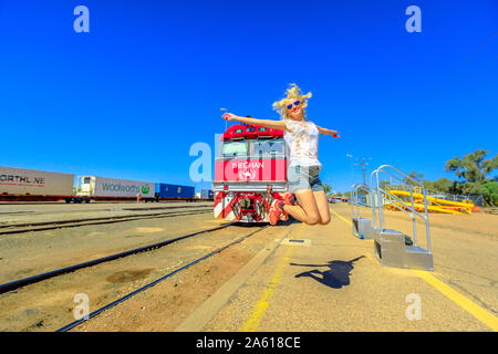 Alice Springs, Northern Territory, Australia - Aug 29, 2019: tourist woman jumping in front of The Ghan iconic luxury passenger train at Alice Springs Stock Photo