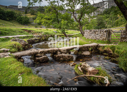 Old stone clapper bridge over Malham Beck with Malham Cove in the background Yorkshire Dales, England, UK. Stock Photo