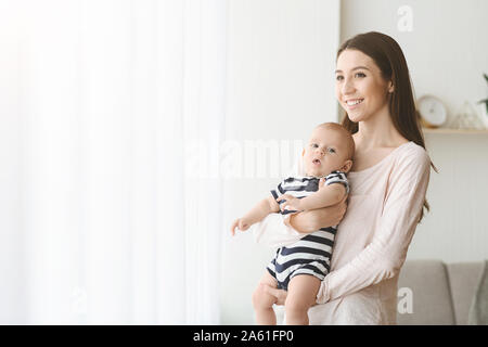 Dreamy woman with little baby on hands standing near window Stock Photo