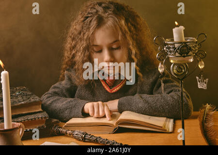 Portrait of a cute sorceress kid with magnificent long brown hair dressed in a navy blue jumper, white shirt and red tie. She is reading a book with s Stock Photo
