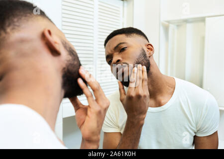 Concentrated african guy looking at his beard at bathroom mirror Stock Photo