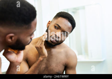 Worried millennial african guy checking his face, looking at mirror Stock Photo