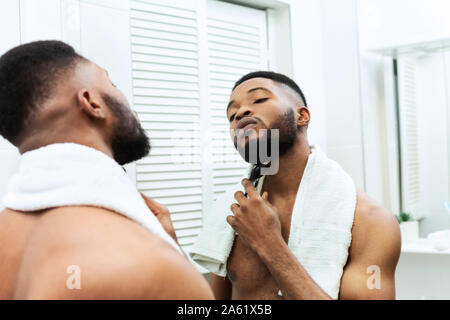 Young man shaving his beard with electric shaver, looking at mirror Stock Photo