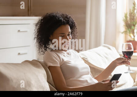 Satisfied young african american woman relaxing on sofa, holding smartphone. Stock Photo