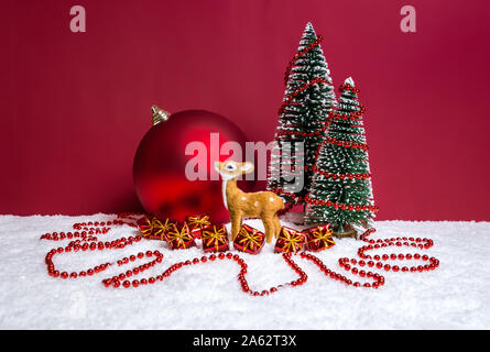 Christmas holiday theme with a miniature cute reindeer, fir trees decorated with red beads, small presents and big Christmas bauble on snow against re Stock Photo