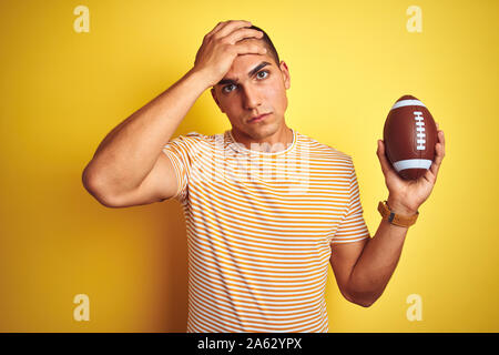 Young rugby player man holding a football ball over yellow isolated background stressed with hand on head, shocked with shame and surprise face, angry Stock Photo