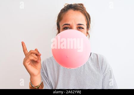 Young redhead woman chewing gum and blowing hair bubble over white isolated background very happy pointing with hand and finger to the side Stock Photo