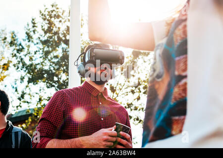 Young bearded male in red shirt wearing virtual reality glasses and big headphones outdoor in the crowd. Smartphone with VR goggles headset. Stock Photo
