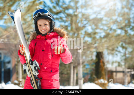 Winter vacation. Girl in goggles and helmet with skis standing outdoors smiling happy showing thumb up close-up blurred background