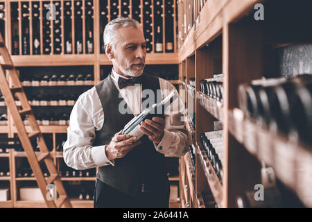 Sommelier Concept. Senior man standing taking wine from cabinet pensive Stock Photo