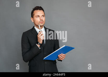 Businessman studio standing isolated on grey with clipboard touchging chin with pen thoughtful Stock Photo