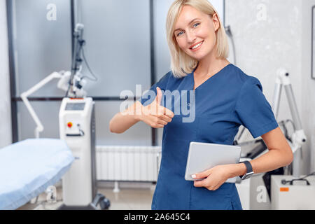 Cosmetology Service. Young doctor standing with digital tablet thumb up smiling cheerful close-up at beauty clinic Stock Photo