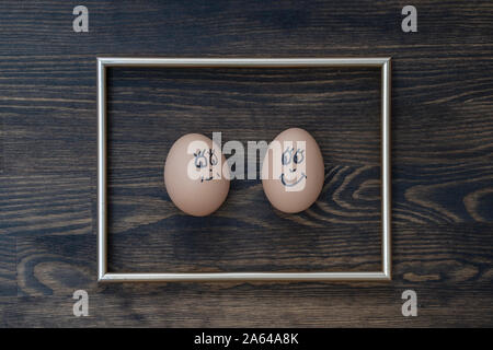 Picture golden frame and two funny eggs smiling on dark wooden wall background, close up. Eggs family emotion face portrait. Couple eggs with happy fa Stock Photo