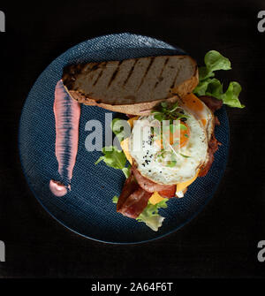 Fried egg bacon cheese lettuce bread roll breakfast served on blue plate Stock Photo