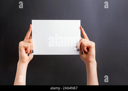 Woman's hands with black nailpolish holding a blank paper against black wall Stock Photo