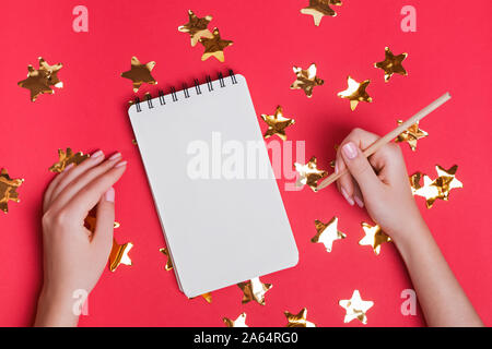 Woman's hands holding pencil near the blank notepad on the red background with star shaped confetti. Stock Photo