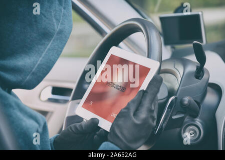 Hooded thief tries to break the car's security systems with tablet. Hacking modern car concept Stock Photo