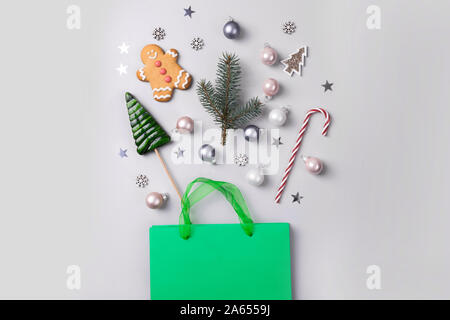 Christmas holiday shopping concept. Green bag with festive purchases, candy cane, treats, decor, glitter confetti on grey background. View from above, Stock Photo
