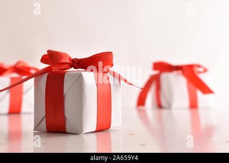 Three white paper gift boxes with red bow on white reflective table and background.. Horizontal composition. Front view. Stock Photo