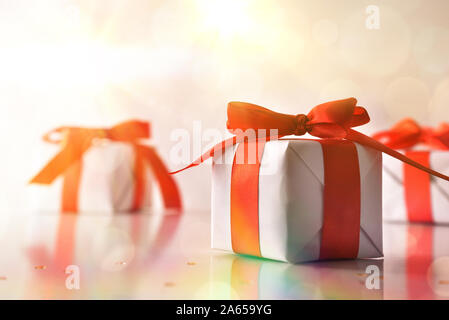 Three white paper gift boxes with red bow on white reflective table and background with warm lights. Horizontal composition. Front view. Stock Photo
