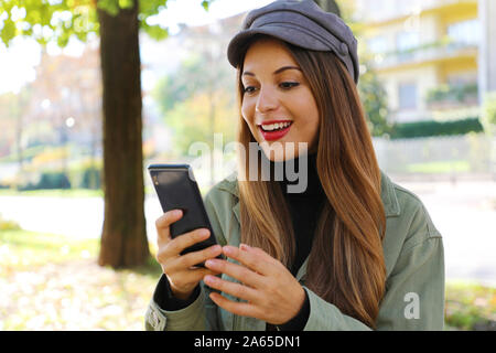 Surprised casual girl wearing hat and polo neck reading good news on smart phone outdoor. Stock Photo