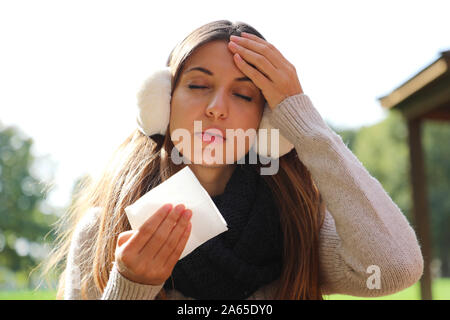 Young woman with earmuffs and scarf suffering migraine headache holding tissue outdoors. Stock Photo
